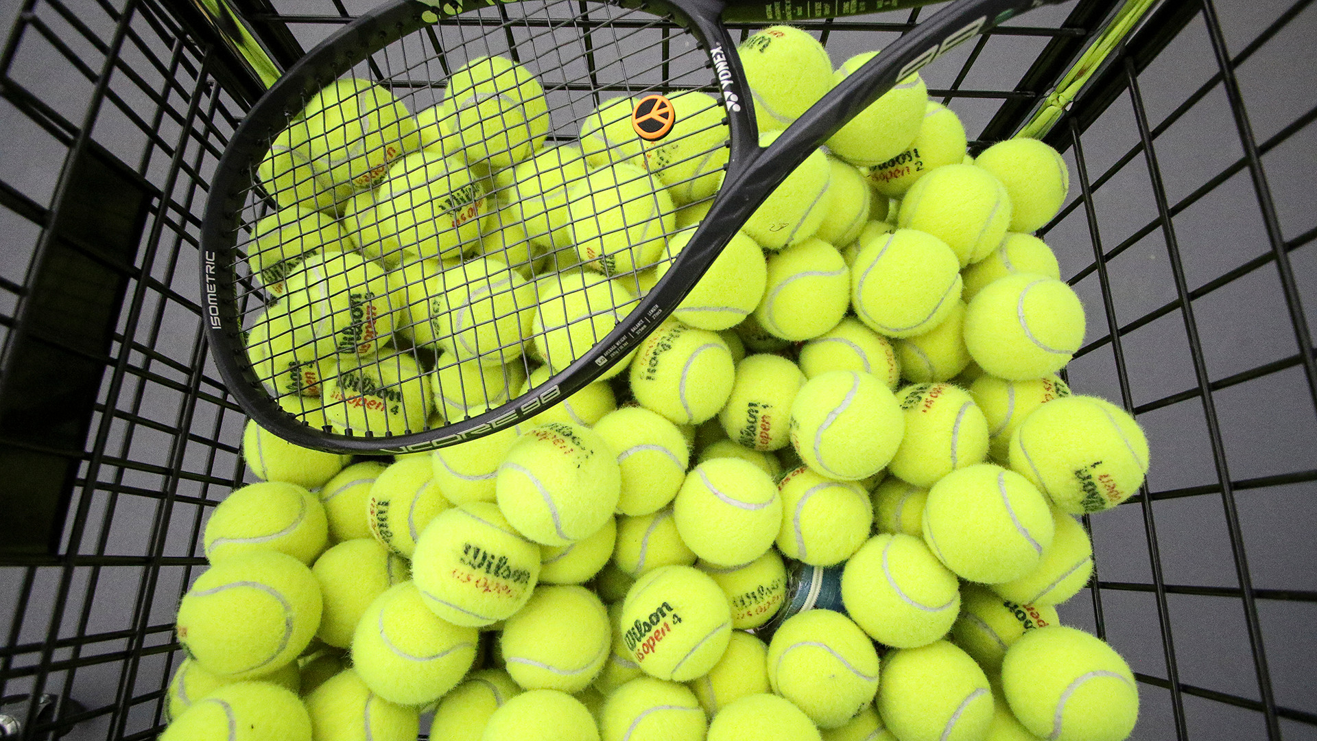 Tennis balls in a wire basket with a racquet sitting on top of it. 