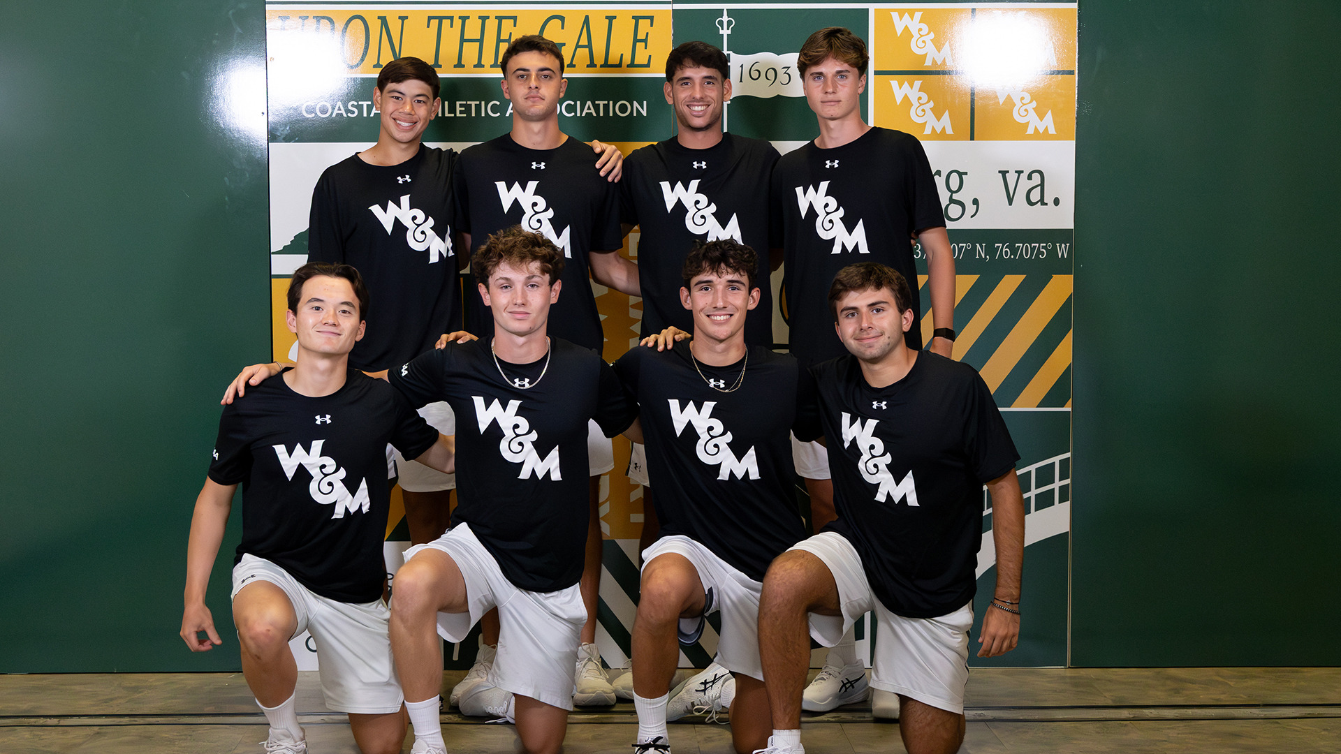 The men's tennis team poses in front of the scrapbook backdrop during media day in two rows with black shirt and white shorts. 