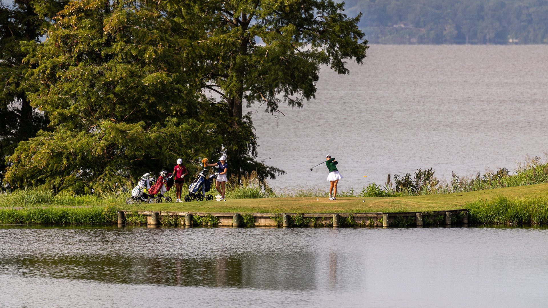 A Tribe player tees off on the 18th hole on the River Course at Kingsmill with water in front of the tee box and the James River behind them. 