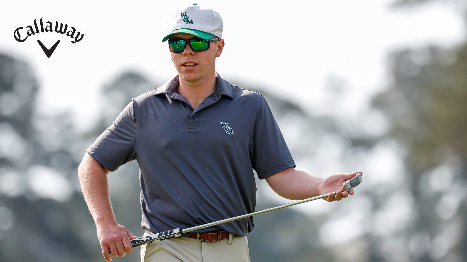 Preston Burton, wearing sunglasses and a white and green W&M hat, walks toward the hole, holding his putter on the practice green at the Golden Horseshoe. 