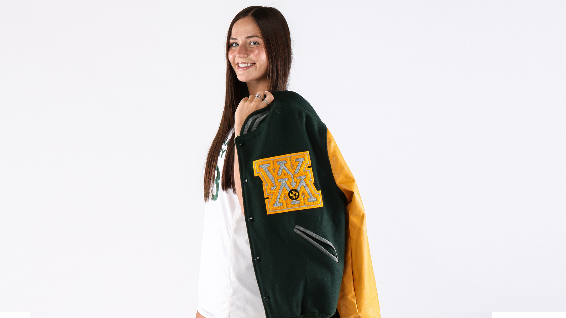 Ivey Crain stands and smiles at side profile holding a women's soccer letterman's jacket and wearing a white soccer uniform during a preseason photo shoot. 