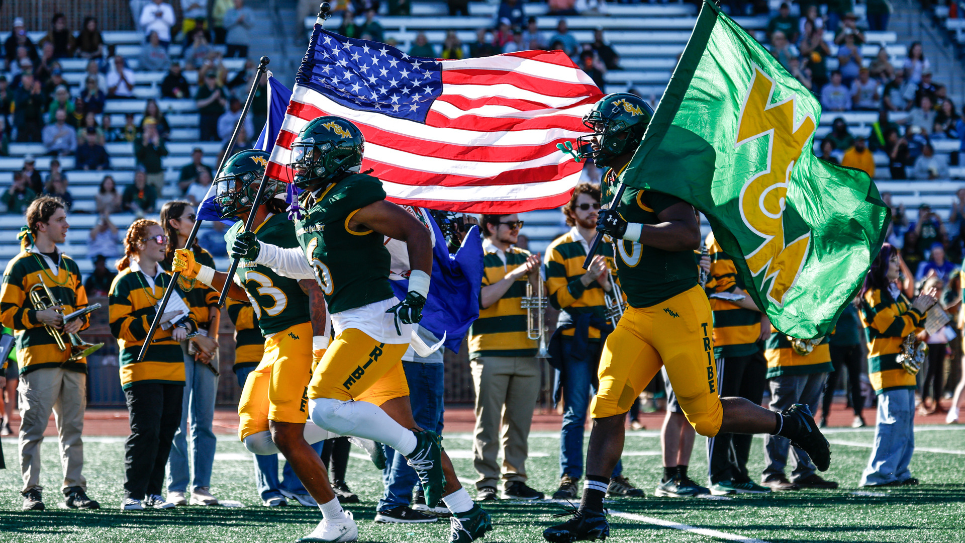 Three W&M football players run on the field holding flags prior to a home game at Zable Stadium