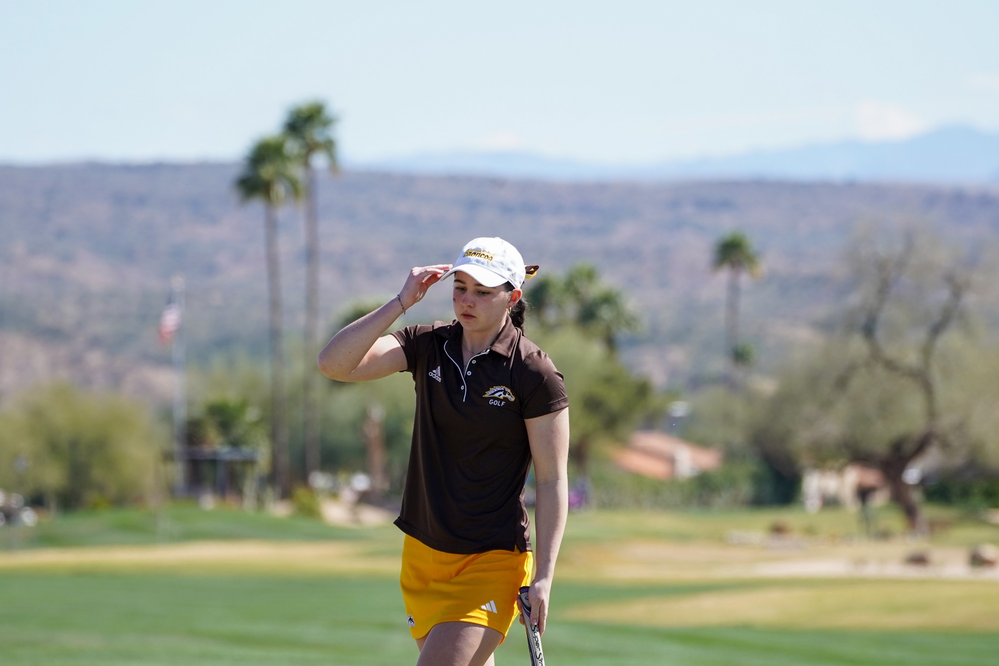 Lea Lemieux holds the brim of her white ball cap with her left hand while looking forward with a golf club in her right hand that is mostly out of frame