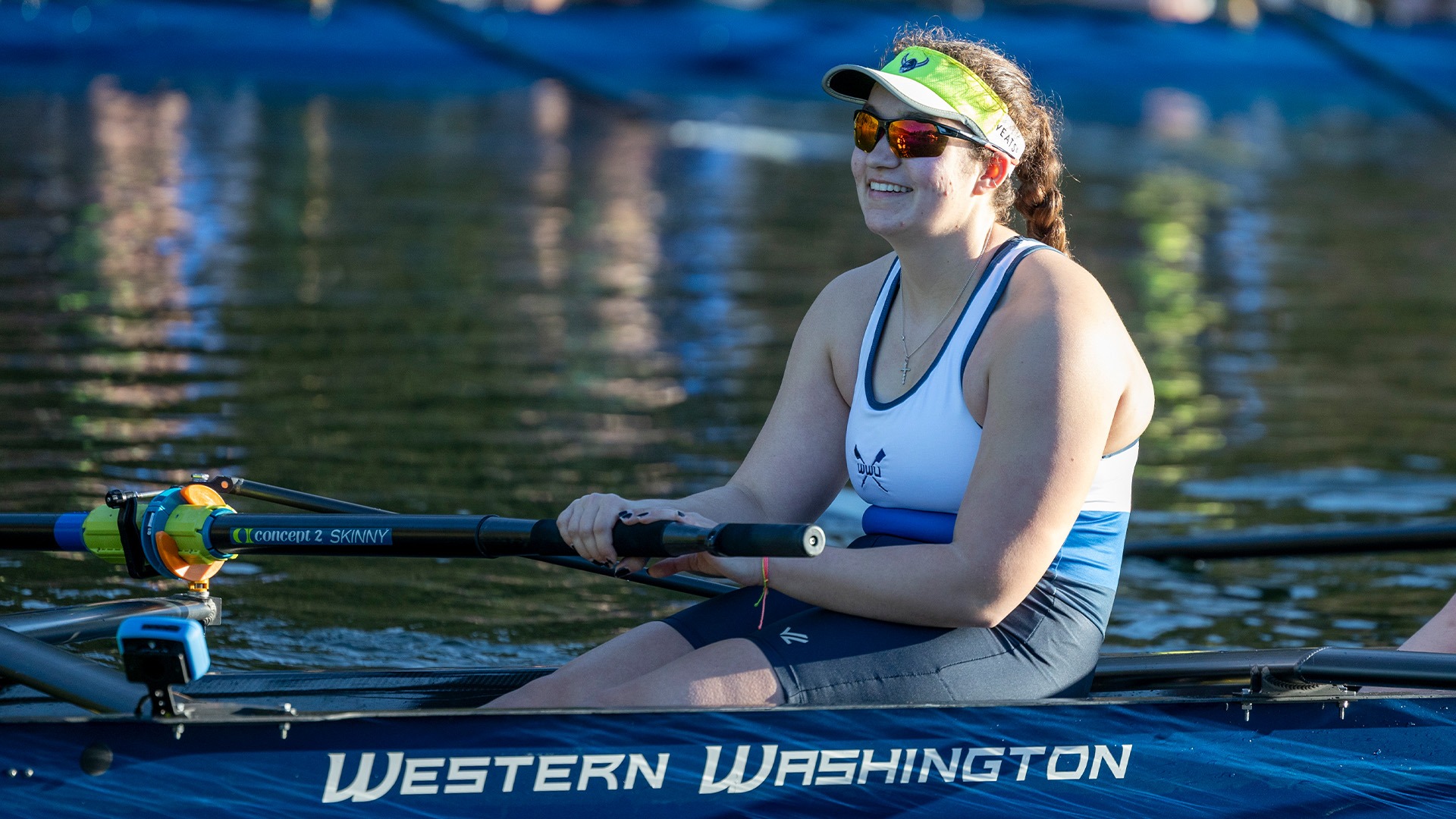 Smiling female rower in a Western Washington uniform, wearing sunglasses and a visor, grips her oar.  The boat's name is visible.