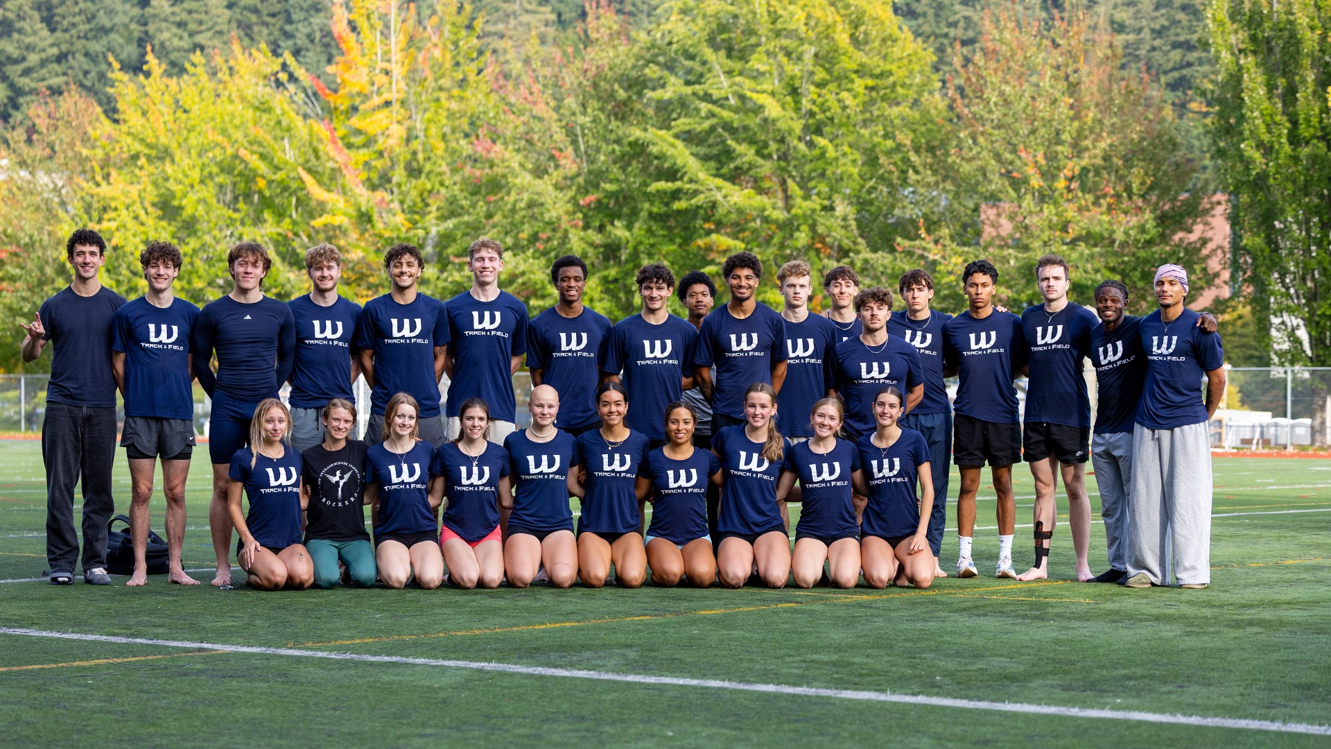 Western Washington University track and field athletes pose together on an outdoor turf field, with one row standing and another kneeling in front. Most wear navy shirts featuring a white Western “W” logo and “Track & Field” lettering. Trees with green and yellow autumn foliage fill the background.