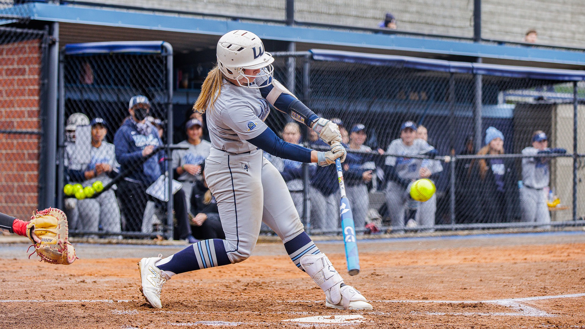 A softball player swings at a pitch, mid-game. The batter is in motion, with the ball in focus.
