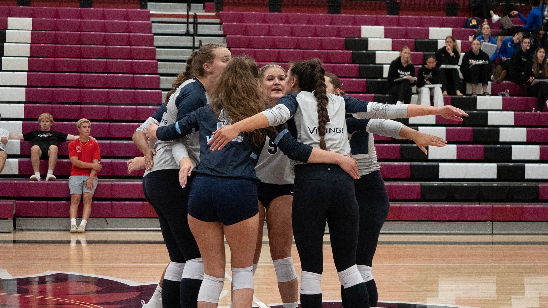 Photo of WWU Volleyball team in gray uniforms in a huddle with bleachers in the background