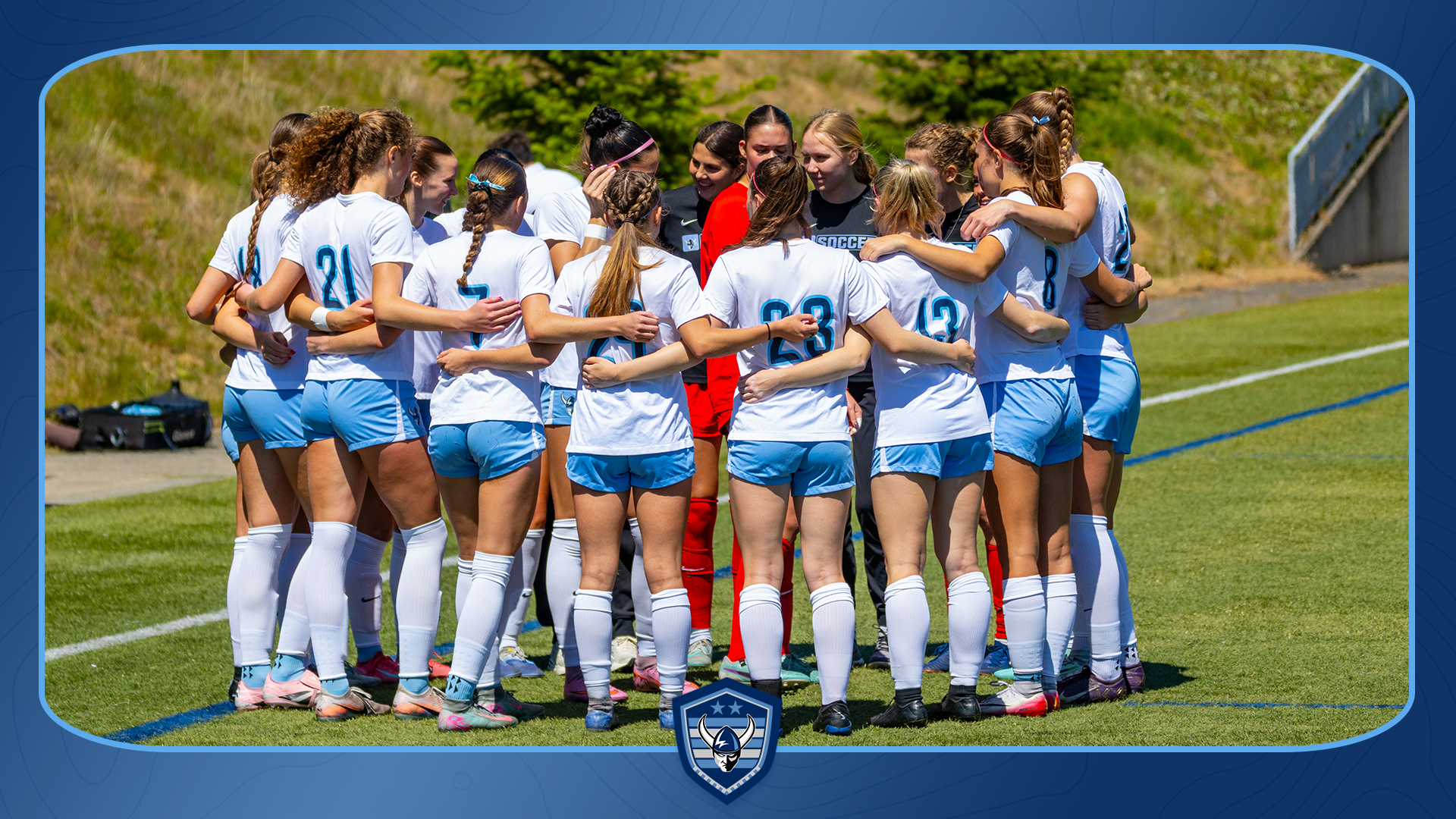 A soccer team in white jerseys and light blue shorts stands in a tight pregame huddle on a sunny outdoor field, with arms around one another near the sideline. A player in a red uniform stands near the center. Trees and a grassy hillside form the background, and the image is framed by a blue graphic border with a team logo at the bottom.