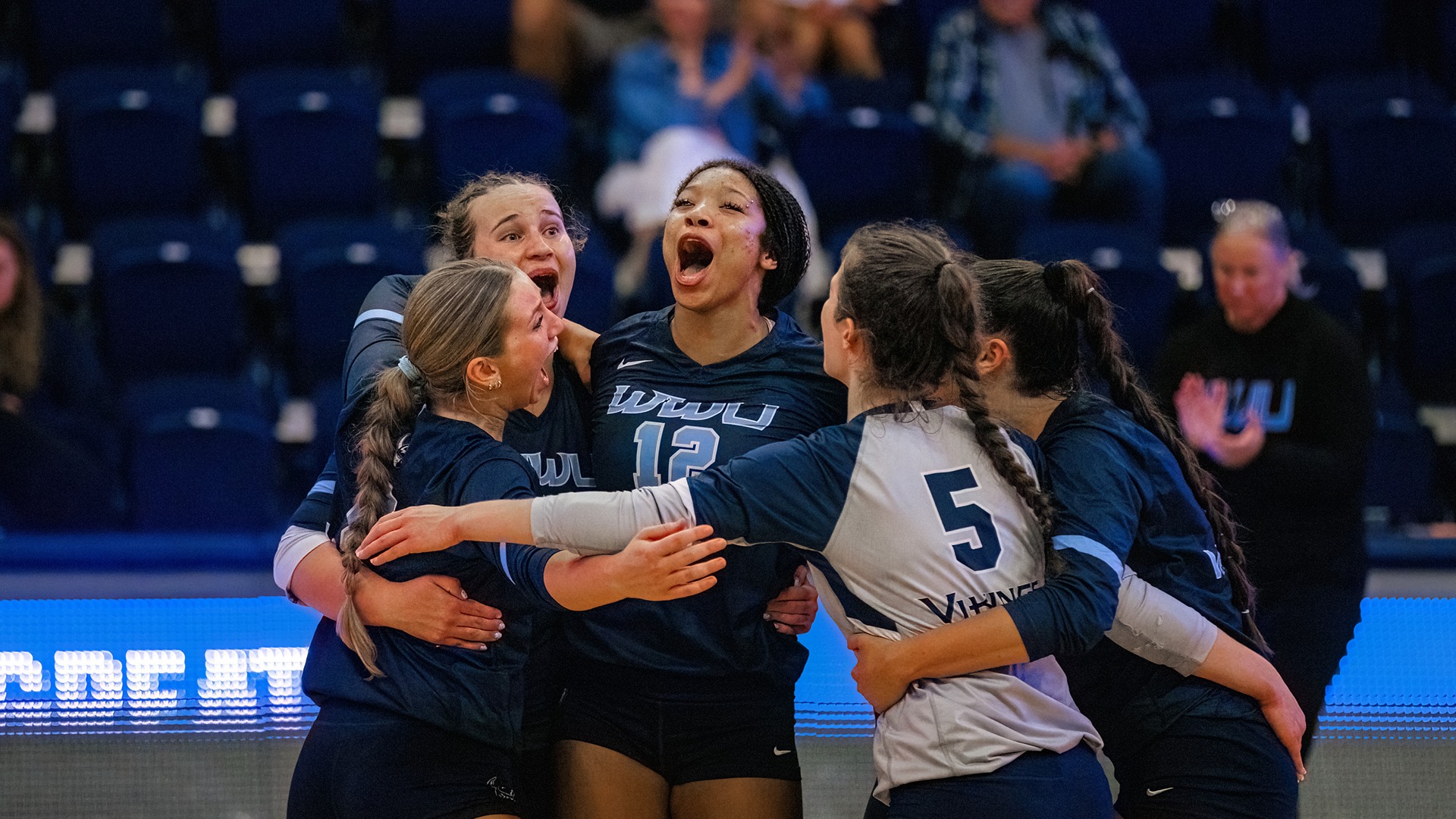 Five volleyball players wearing navy and white uniforms gather in a tight huddle on an indoor court during a match. The athletes have their arms around one another and appear to be celebrating a successful play. The player at the center wears a jersey displaying “WWU” and the number 12, while another player wears a white libero jersey with the number 5. Blue arena seating and spectators are visible in the softly blurred background, drawing attention to the team’s on-court unity and energy.