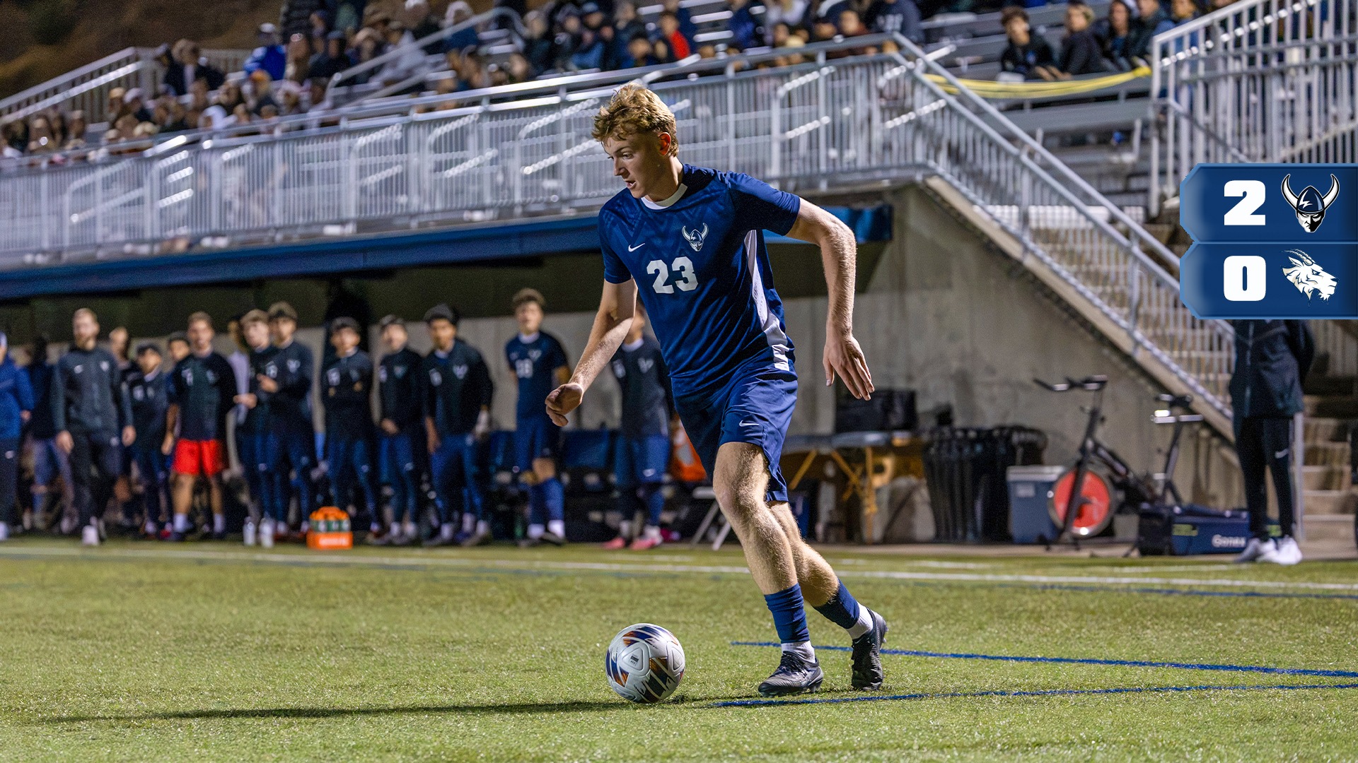 A soccer player in a blue uniform with the number 23 controls the ball near the touchline during a night game on a turf field. Team members stand along the sideline behind the player, while spectators fill the stadium bleachers in the background. Stadium lights illuminate the field, and a scoreboard graphic on the right side of the image shows a 2-0 score with team logos.