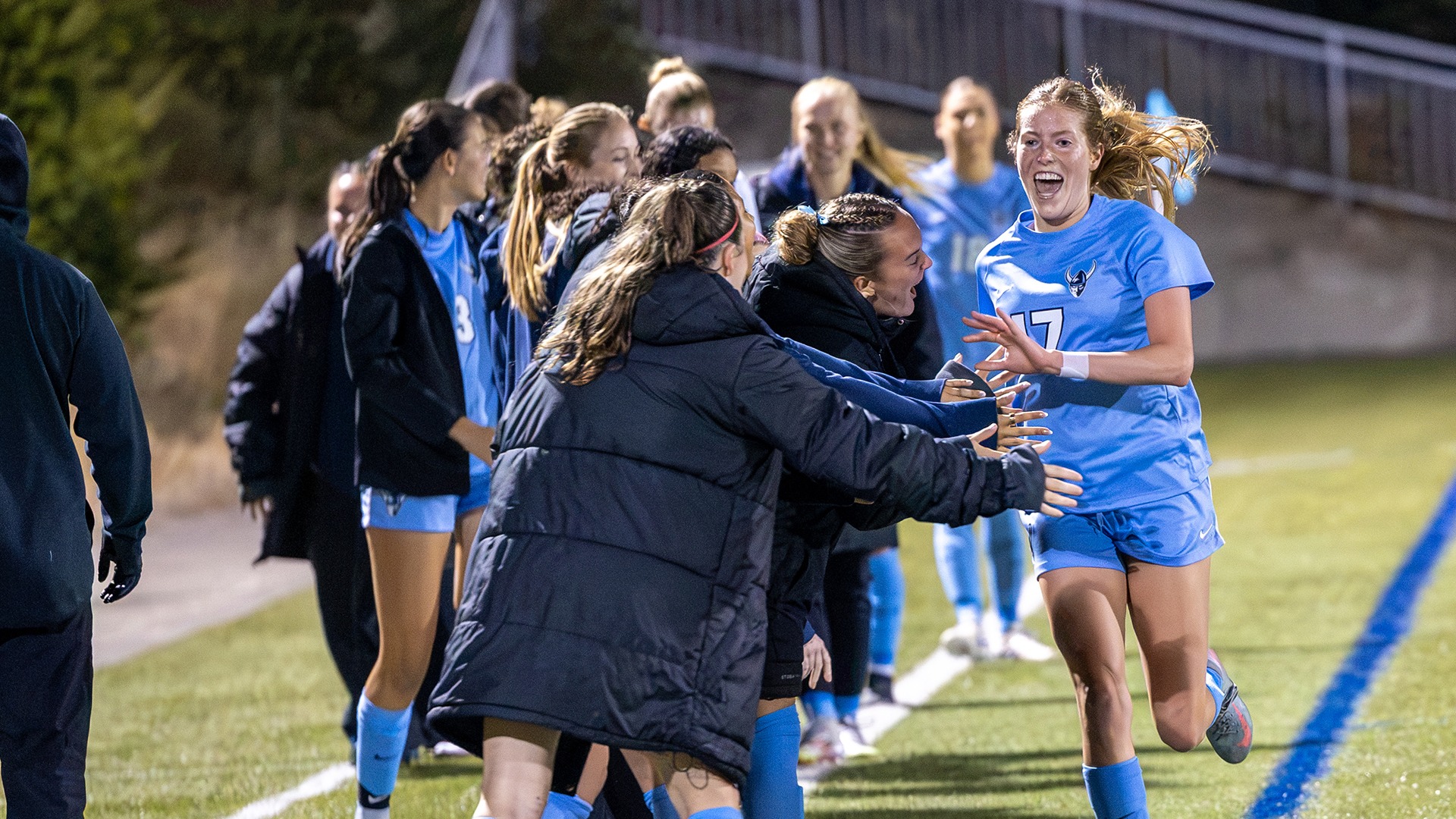 A soccer player wearing a light blue jersey with the number 17 runs down the sideline while teammates in matching uniforms and dark jackets reach out from the bench area. The scene takes place on a lit soccer field at night, with players gathered near the touchline and stadium structures visible in the background.