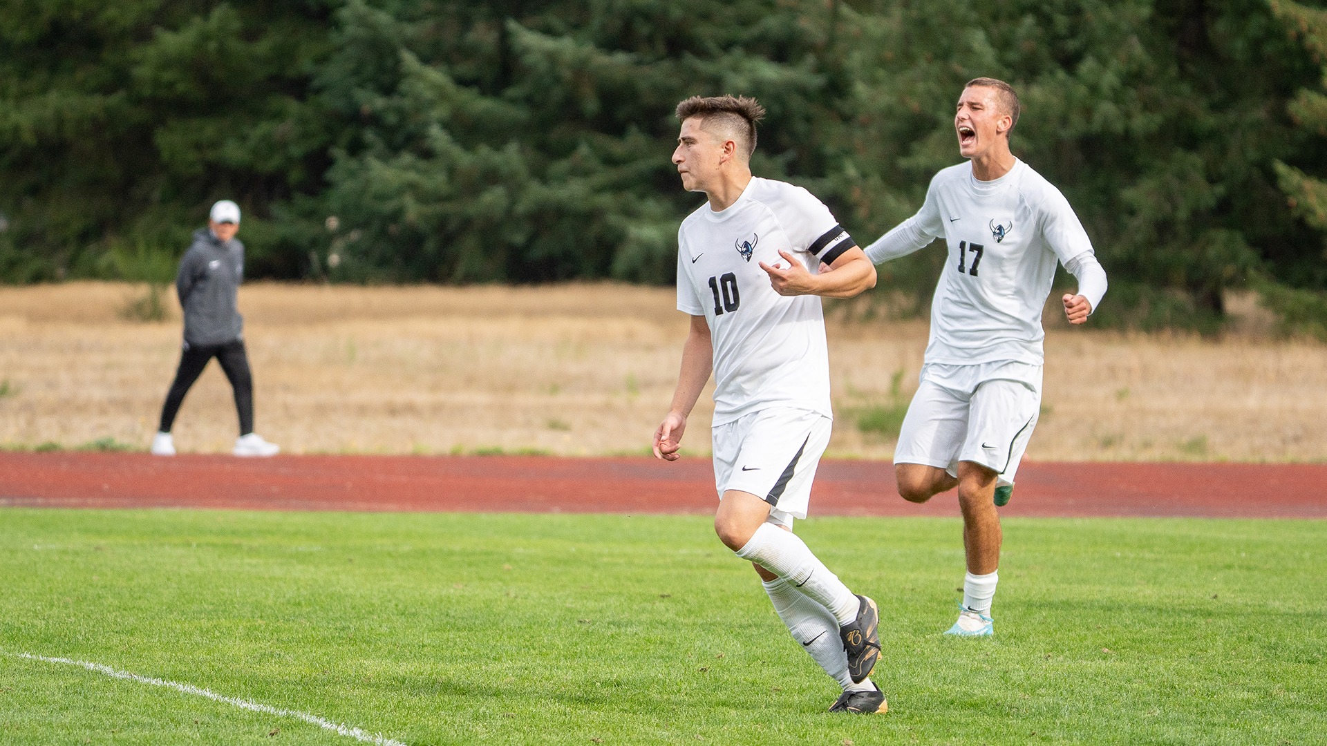 Two Western Washington University men’s soccer players in white uniforms run across a green field. In the foreground, No. 10 wears a black captain’s armband and looks to his left as he runs. Behind him, No. 17 runs with his mouth open and arms spread, appearing to celebrate. A person in gray stands near the track in the background, with trees beyond the field.