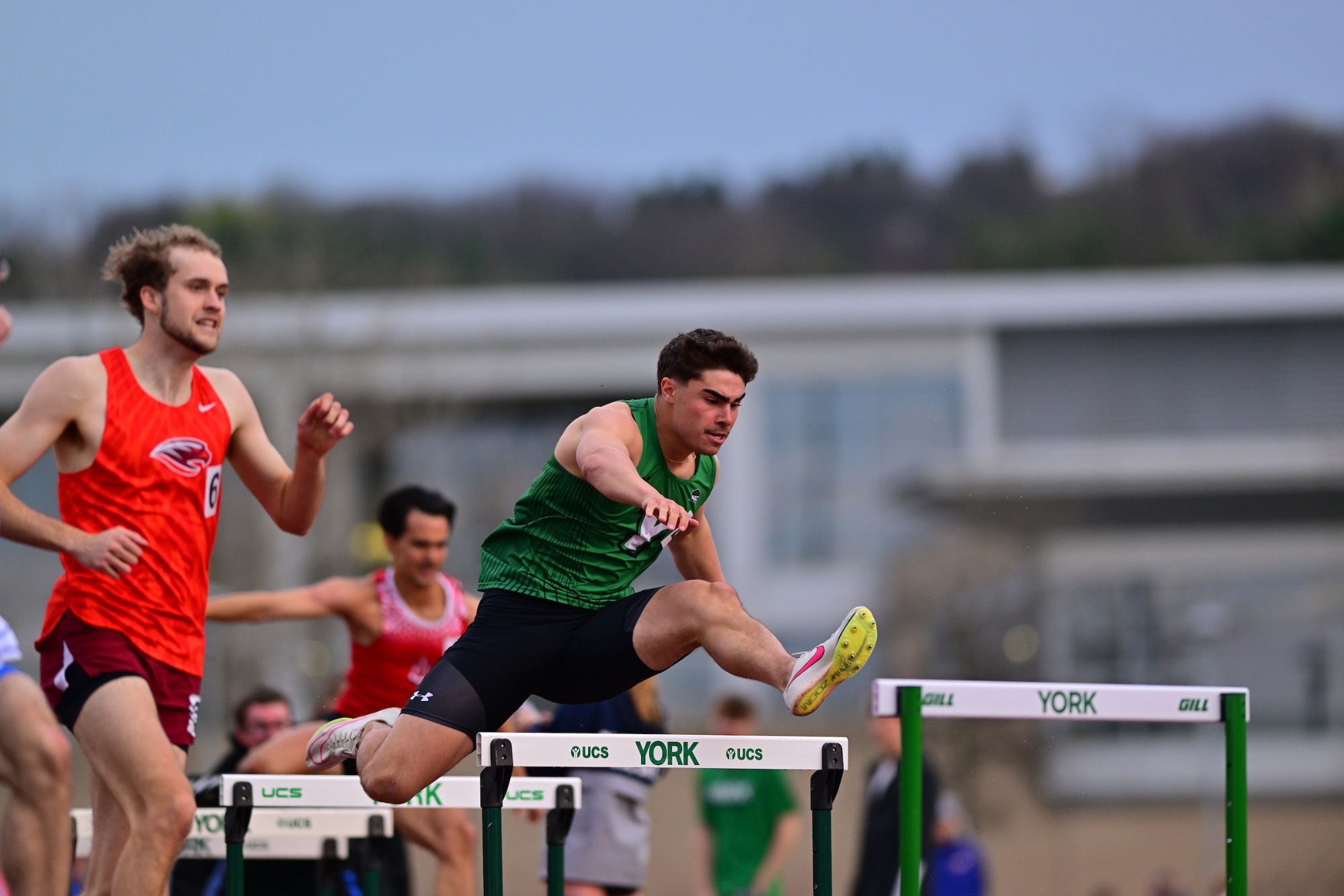 male in green uniform jumping over hurdle