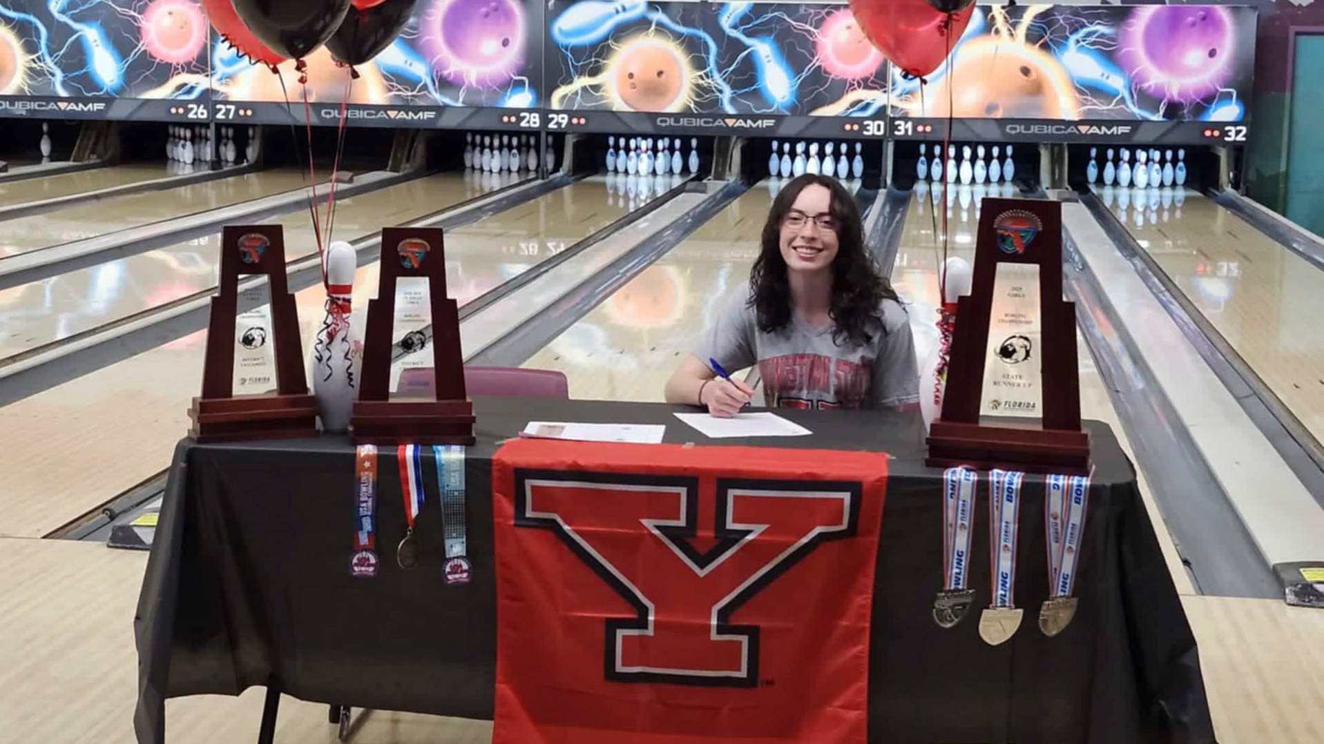 Malayna Calo sits at a table with trophies and medals in front of bowling lanes as she signs her commitment to Youngstown State.