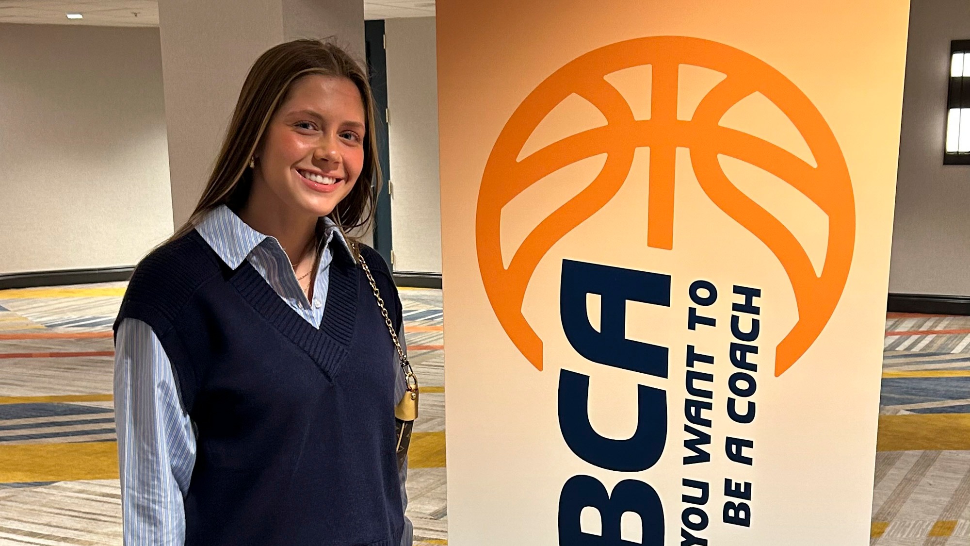 Casey Santoro stands to the left of a vertical sign for the WBCA So You Want To Be A Coach program